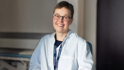 Ihotautilääkäri Kati Pummi istuu vastaanottohuoneessa lääkärintakki yllään ja hymyilee.