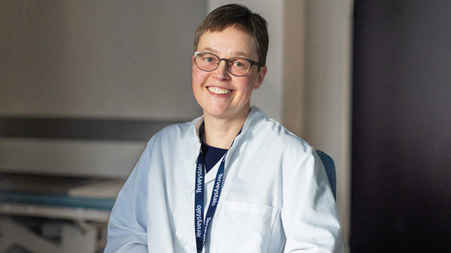 Ihotautilääkäri Kati Pummi istuu vastaanottohuoneessa lääkärintakki yllään ja hymyilee.