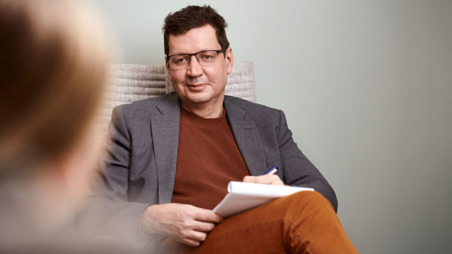 Psykiatri Sami Leppämäki istuu vastaanotolla nojatuolissa käsissään paperinippu ja hymyilee etualalla istuvalle potilaalle.