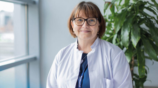 Yleislääkäri Marja-Leena Hyypiä seisoo aulatilassa ikkunan vieressä ja hymyilee. Taustalla korkea viherkasvi. 
