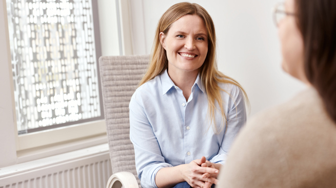 Kauluspaitaan pukeutunut psykiatri istuu tuolissa ja hymyilee etualalla istuvalle potilaalle.