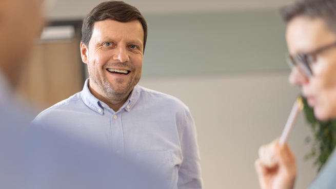 Psykiatri hymyilee kollegalle iloisesti. Etualalla kuvassa toinen psykiatri mietteliäänä kynän kanssa.