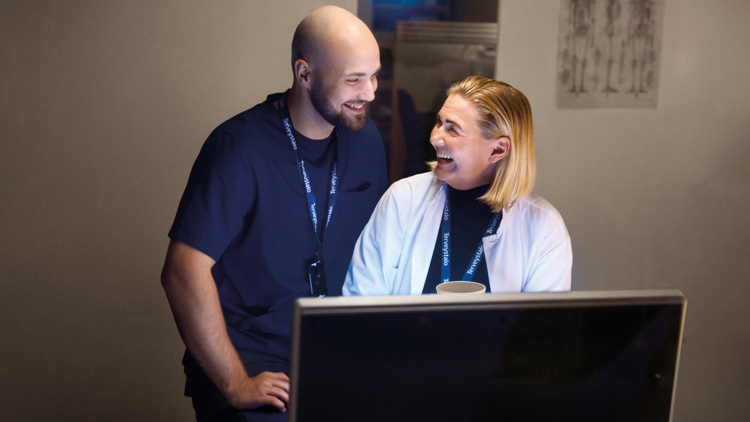 Röntgenhoitaja ja lääkäri juttelevat iloisesti tietokoneen takana ja katsovat toisiaan hymyillen.